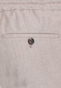 Primo piano di un tessuto beige chiaro che mostra un singolo bottone nero e una tasca orizzontale con un sottile dettaglio di cucitura.