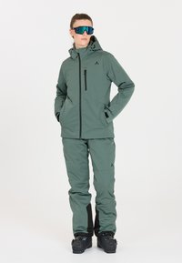 Grüne Skijacke und -hose aus wasserabweisendem Material, mit einer Reißverschlusstasche und schwarzen Akzenten, sowie reflektierenden Sonnenbrillen.