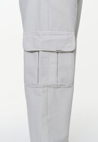 Pantaloni cargo gri deschis, cu un buzunar lateral cu capac, realizați dintr-un material durabil, cu detalii de coasere vizibile.