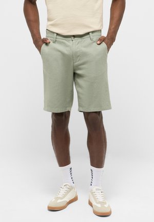 Man wearing light green knee-length shorts with hands in pockets, white crew socks with branding, and cream sneakers with gum soles.