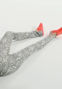 Benen met grijze leggings met slangenprint en feloranje instapschoenen op een effen lichte achtergrond.
