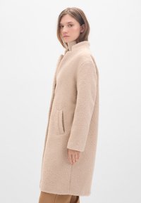Beige, strukturiertem Mantel mit hohem Kragen, langen Ärmeln und einer Seitentasche. Weicher, plüschiger Stoff mit lockerer Passform und minimalistischem Design.