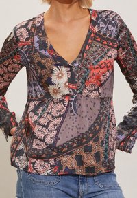 Kvinna i blus med lång ärm och V-ringning med flerfärgade blommiga och paisleymönster, matchat med blå jeans.