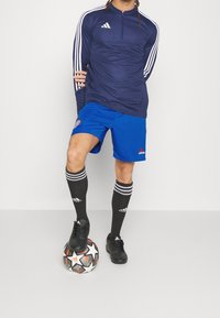 Muž v modrém sportovním oblečení, který balancuje nohu na fotbalovém míči, nosí černé ponožky Adidas a kopačky s bílými pruhy na rukávech.