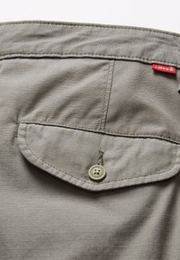 Camicia di tessuto grigio con una sola tasca a busta, dotata di un bottone grigio chiaro, dettagli cuciti e una piccola etichetta rossa del marchio. Superficie strutturata.