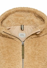 Fleecegevoerde jas in lichtbruin, met een ritsluiting, capuchon en een binnenvoering van gewatteerd materiaal met een label. Zachte, harige buitenkant.