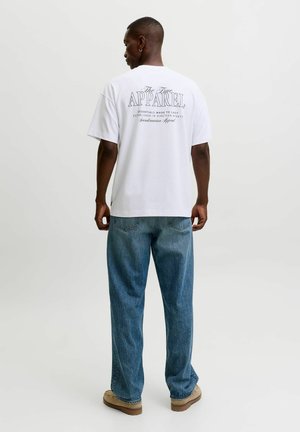 Mand iført hvid t-shirt med tekst på bagsiden og blå jeans, stående med fronten væk mod en enkel lys baggrund.