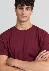 T-shirt in cotone borgogna con maniche corte, design a girocollo, stile attillato, texture liscia, indossata da una persona con le braccia incrociate.