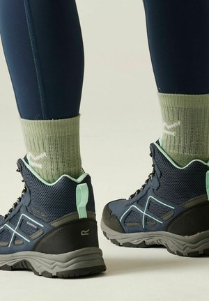 Persoon die zwarte en muntgroene wandelschoenen draagt met lichtgroene sokken en zwarte legging, staand op een lichte achtergrond.