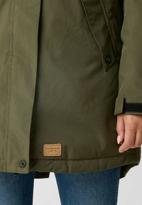 Olijfgroene parka met een gestikte zoom, drukknoopsluitingen en een tan leren merkpatch aan de onderkant. De stof heeft een gladde textuur.