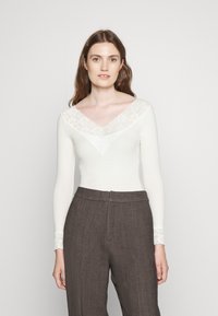 Pitkähiha bodysuit kerma värissä, jossa on pitsinen V-mallinen kaulus ja pitsikoristeet hihoissa, yhdistetty korkeavyötäröisten, tekstitettyjen ruskeiden housujen kanssa.