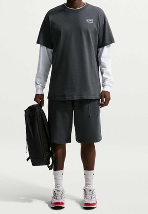 Młody mężczyzna w czarnych szortach, czarnej koszulce Nike na białej koszuli z długim rękawem, białych skarpetkach Nike, butach sportowych, trzymający czarny plecak w jednej ręce.