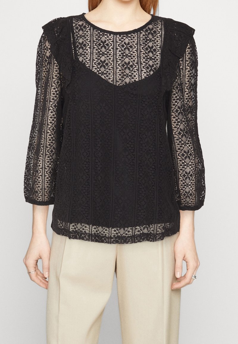 Blusa di pizzo nero con maniche a tre quarti a volant, che presenta motivi intricati e un orlo scallop. Indossata sopra pantaloni beige.