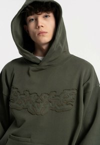 Oliwkowa bluza z dużym kapturem, z teksturowanym, abstrakcyjnym wzorem w ciemniejszej zieleni na klatce piersiowej oraz kieszenią z przodu.
