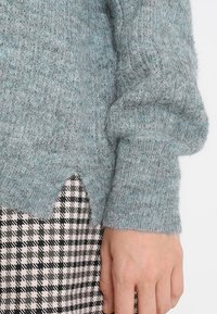 Pull bleu clair texturé avec une apparence douce et un ourlet en zigzag, associé à un motif pied-de-poule noir et blanc en bas.