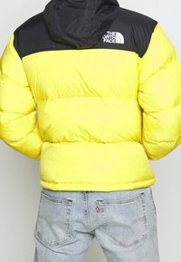 Żółta kurtka puchowa z czarnymi ramionami i kapturem, z gładką fakturą i logiem na górnej części pleców, w połączeniu z jasnoniebieskimi dżinsami.