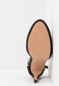 Chaussure à talon haut en daim noir avec un bout pointu, un talon fin et une semelle lisse marron clair. Présente de fines lanières noires autour de la cheville.