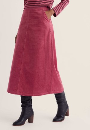 Woman wearing a pink corduroy midi skirt with side pockets and black heeled boots, paired with a pink and purple striped long-sleeve top.