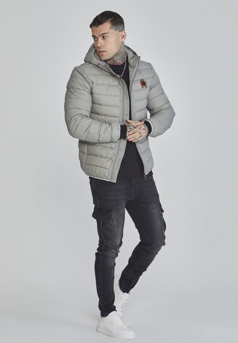 Graue Steppjacke mit Kapuze, ausgestattet mit einem gesteppten Design und einem kleinen gestickten Patch. Getragen über einem schwarzen gerippten Pullover und Cargo-Hosen.