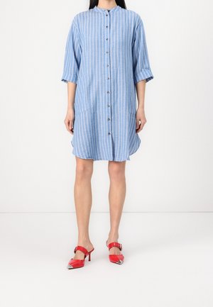 Kobieta ubrana w niebiesko-białą sukienkę w paski sięgającą kolan, zapinaną na guziki, z rękawami do połowy przedramienia, oraz czerwone buty na obcasie z noskiem w szpic.