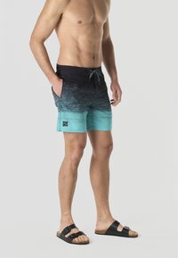 Uomo senza maglietta che indossa pantaloncini da bagno sfumati neri e turchesi con coulisse, mani nelle tasche e sandali neri senza lacci, su sfondo semplice.