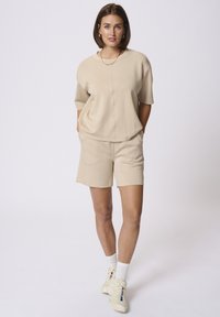 Sweatshirt beige à manches courtes et Short assorti, en tissu tricoté, coupe décontractée, col rond, avec de subtils détails de couture. Baskets blanches aux pieds.