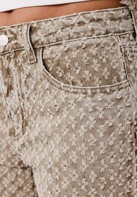 Los pantalones de mezclilla beige presentan un patrón texturizado con motivos en relieve. Incluyen un diseño clásico de bolsillos y un cierre frontal con botón.