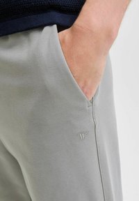 Mano appoggiata nella tasca anteriore di pantaloni grigio chiaro con un piccolo logo ricamato vicino alla cucitura della tasca.