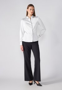 Camicia bianca in satin con bottoni, piping nero, maniche lunghe e colletto, abbinata a pantaloni neri dritti e tacchi neri.