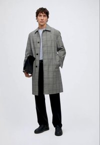 Homme portant un manteau à carreaux gris, une chemise gris clair, un pantalon noir et des chaussures noires, tenant un objet noir plié dans une main.