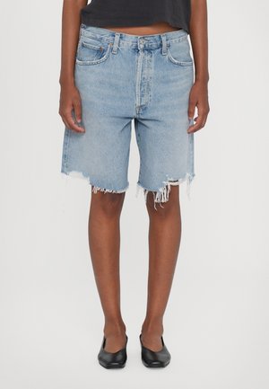 Persoon draagt lichtblauwe versleten denim shorts, zwarte platte schoenen en een zwart cropped top, staand tegen een effen achtergrond.