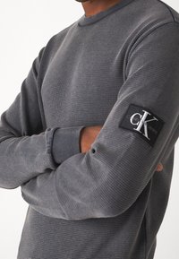 Sweatshirt gris en maille gaufrée avec poignets et encolure côtelés. Présente une étiquette rectangulaire noire avec logo blanc sur la manche gauche.