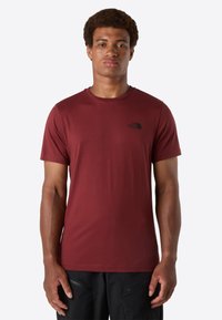 Mann steht mit Blick nach vorne und trägt ein einfarbiges weinrotes T-Shirt mit kurzen Ärmeln, das ein kleines schwarzes Logo auf der linken Brust hat, sowie schwarze Hosen.
