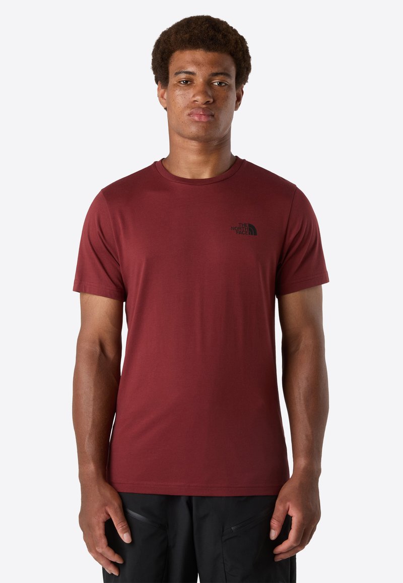 Mann steht mit Blick nach vorne und trägt ein einfarbiges weinrotes T-Shirt mit kurzen Ärmeln, das ein kleines schwarzes Logo auf der linken Brust hat, sowie schwarze Hosen.