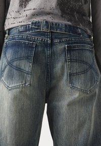 Denim jeans with a faded blue color, featuring two back pockets with decorative stitching, and a metal buckle at the waistband.