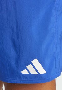 Pantalones cortos azules de tela suave presentan un logo blanco de Adidas con tres rayas diagonales en la parte inferior izquierda. Se aprecia un detalle de costura limpia.