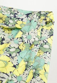 Tissu avec un motif floral dense présentant des marguerites jaunes, blanches et vertes sur un fond sombre, montrant un détail d'angle plié.