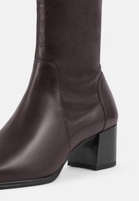 Bordeaux rode leren kniehoge laars met een vierkante hak, een gladde textuur en stikseldetail, met een strak en modern ontwerp.