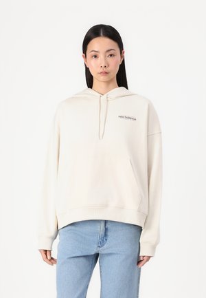 Crème-kleurige hoodie met een voorzak, trekkoordcapuchon en geborduurd 'new balance' logo. Gecombineerd met lichtblauwe jeans.