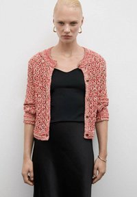 Gehaakte koraalkleurige cardigan met een ronde halslijn, lange mouwen, knoopsluiting en open brei-textuur. Draagover een zwarte top.