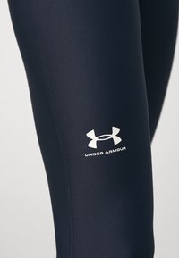 Granatowe legginsy wykonane z elastycznego materiału o gładkiej fakturze. Posiadają biały logo Under Armour na dolnej części nogawki oraz płaskie szwy.