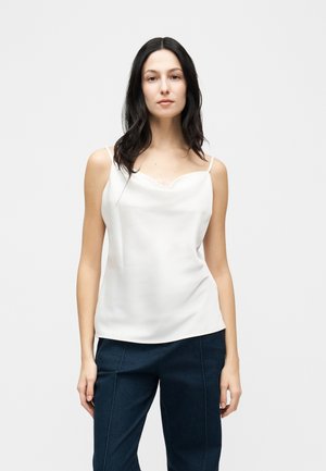 Kobieta o długich ciemnych włosach, ubrana w białą, bezrękawnikową bluzkę i ciemnoniebieskie spodnie, stojąca na tle jednolitego jasnego tła.