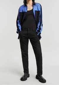 Chemise bleu et noire à blocs de couleur, boutonnée, avec deux poches poitrine. Un pantalon noir ajusté et des bottes noires épaisses complètent la tenue.