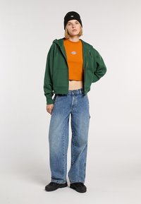 Sudadera verde con cremallera, camiseta corta naranja con logo, vaqueros anchos de color azul claro, gorro negro y zapatos negros. Conjunto casual en capas.
