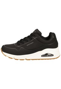 Chaussures de sport noires avec une tige synthétique lisse et des accents perforés. Elles présentent une semelle intermédiaire blanche et une semelle extérieure en caoutchouc gomme avec un coussin d'air visible.