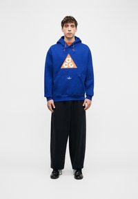 Νεαρός άνδρας όρθιος, φορώντας μπλε φούτερ με κουκούλα και πορτοκαλί λογότυπο σε σχήμα τριγώνου, φαρδιά μαύρα παντελόνια και μαύρα και λευκά αθλητικά παπούτσια, μπροστά από ένα απλό φόντο.