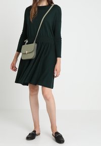 Knarvlång mörkgrön klänning med rund halsringning och långa ärmar, med en figursydd midja och utsvängd kjol; matchad med en liten grön handväska.