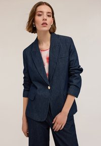 Denimowa marynarka w ciemnym granacie, zapinana na jeden guzik, z dwoma przednimi kieszeniami, z usztywnionymi ramionami, o półluźnym kroju, zestawiona z jasnym, wzorzystym topem.