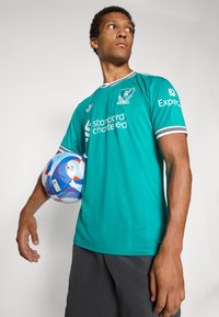 Turkis fotballtrøye med svart og hvitstripet krage, med Liverpool FC-logo og teksten "Standard Chartered" påtrykt. Spilleren holder en flerfarget ball.