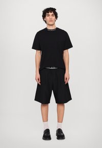 Melna T-kreka ar logotipa apkakli, saskaņota ar brīvām melnām šortiem. Modelis valkā baltas zeķes un melnas kurpes. Minimālistisks dizains un monohromas krāsas.